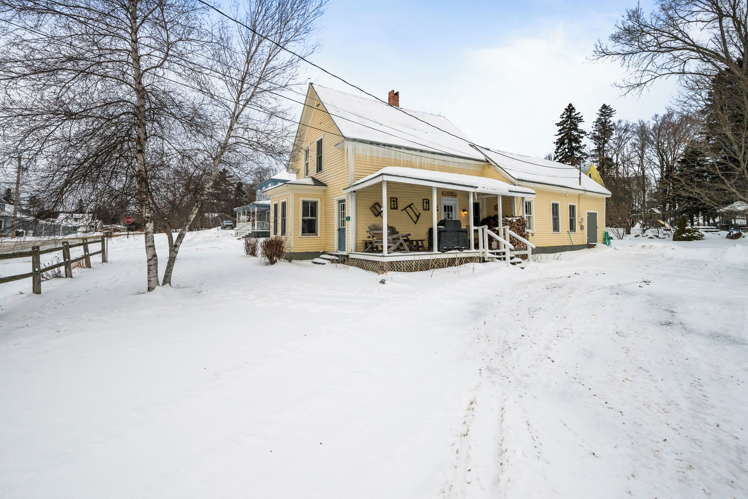 A yellow house with a porch and a chimney, surrounded by snow and trees.