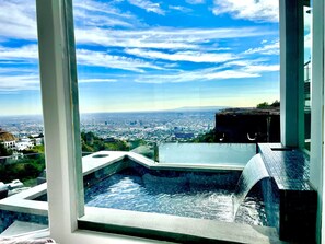 Una habitación de hotel moderna con un ventanal grande que ofrece vistas a un paisaje urbano y una pequeña fuente de agua en el interior.