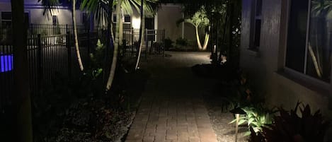 A paved pathway leading to a building with a gated entrance, surrounded by palm trees and plants.