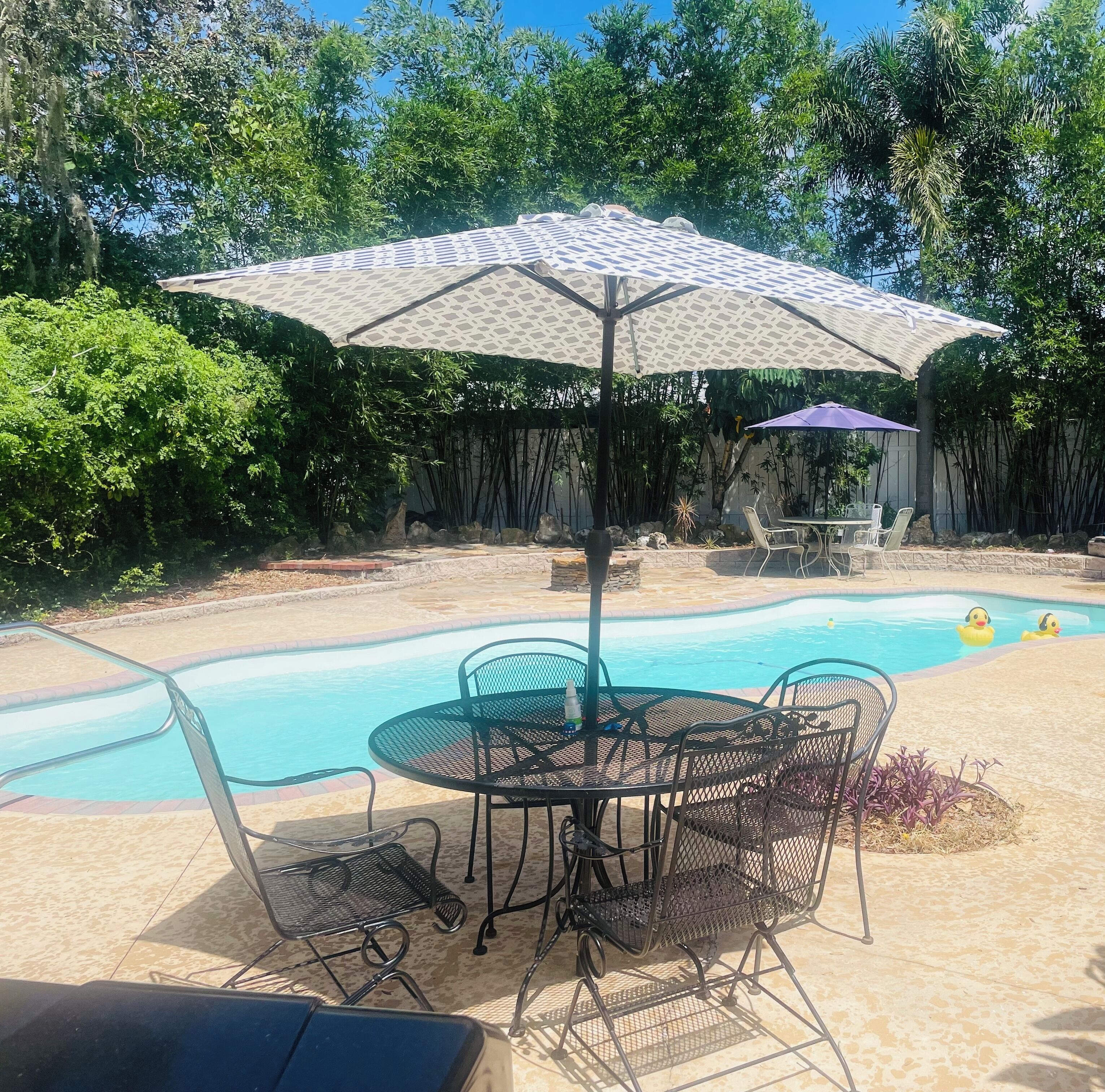 Un patio trasero con piscina, una mesa con sillas y dos sombrillas.