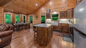 A wooden cabin interior with a dining area, kitchen, and large windows. - Knotty Dreams - Luxury Awaits, Hot Tub, Party Porch (Mineral Bluff)