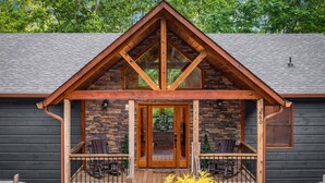 A log cabin with a stone facade, a wooden porch with seating, and a large window above the entrance. - Knotty Dreams - Luxury Awaits, Hot Tub, Party Porch (Mineral Bluff)
