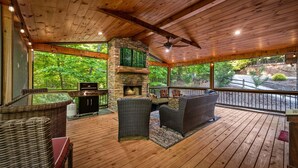A wooden deck with wicker furniture, a stone fireplace, and a view of the forest. - Knotty Dreams - Luxury Awaits, Hot Tub, Party Porch (Mineral Bluff)