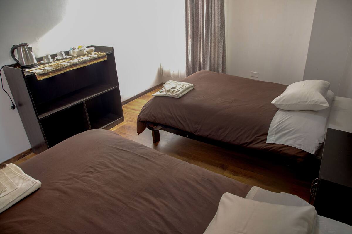 Habitación de hotel con dos camas, una mesita pequeña con una tetera, tazas y una bandeja, y un estante con ropa doblada.