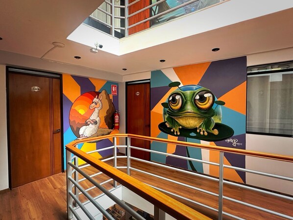 A modern hotel lobby with a wooden floor, a metal railing, and a colorful mural featuring a llama and a frog. - MAGIC PACKER HOSTEL (Cusco)