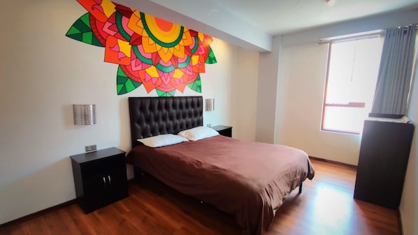 A bedroom with a tufted headboard, a bed with a brown bedspread, a colorful wall decoration, a nightstand, and a window with curtains. - MAGIC PACKER HOSTEL (Cusco)
