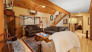 A cozy living room with two sofas, a coffee table, and a fireplace. There is a wooden staircase and a large window with a view of trees. - Riverfront Cabin with Hot Tub and Game Room (Boone)