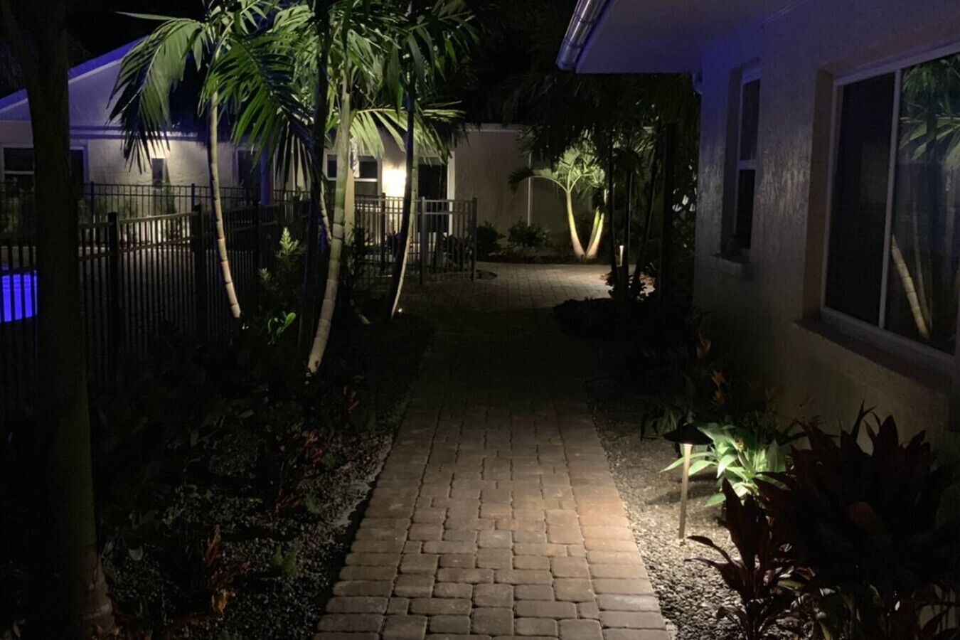 A paved pathway leading to a building with a gated entrance, surrounded by palm trees and plants.
