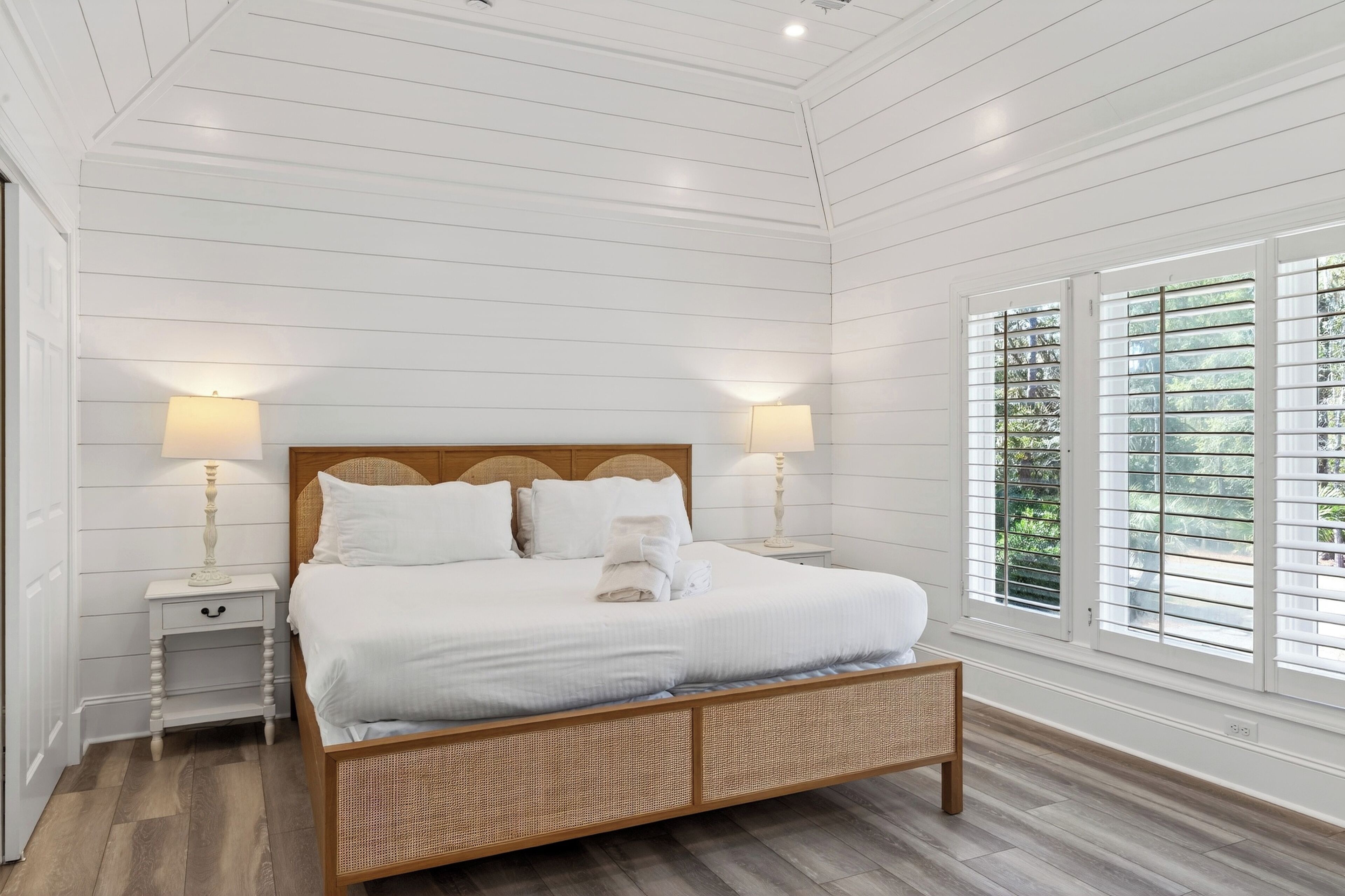 A bedroom with a wooden headboard, white bedding, two bedside tables with lamps, and large windows with blinds.