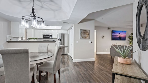 A modern kitchen with white cabinets, a dining area with a round table and chairs, and a living room with a TV and a large plant. - Beautiful Newly Remodeled 2-bdrm Biltmore Condo! (Phoenix)