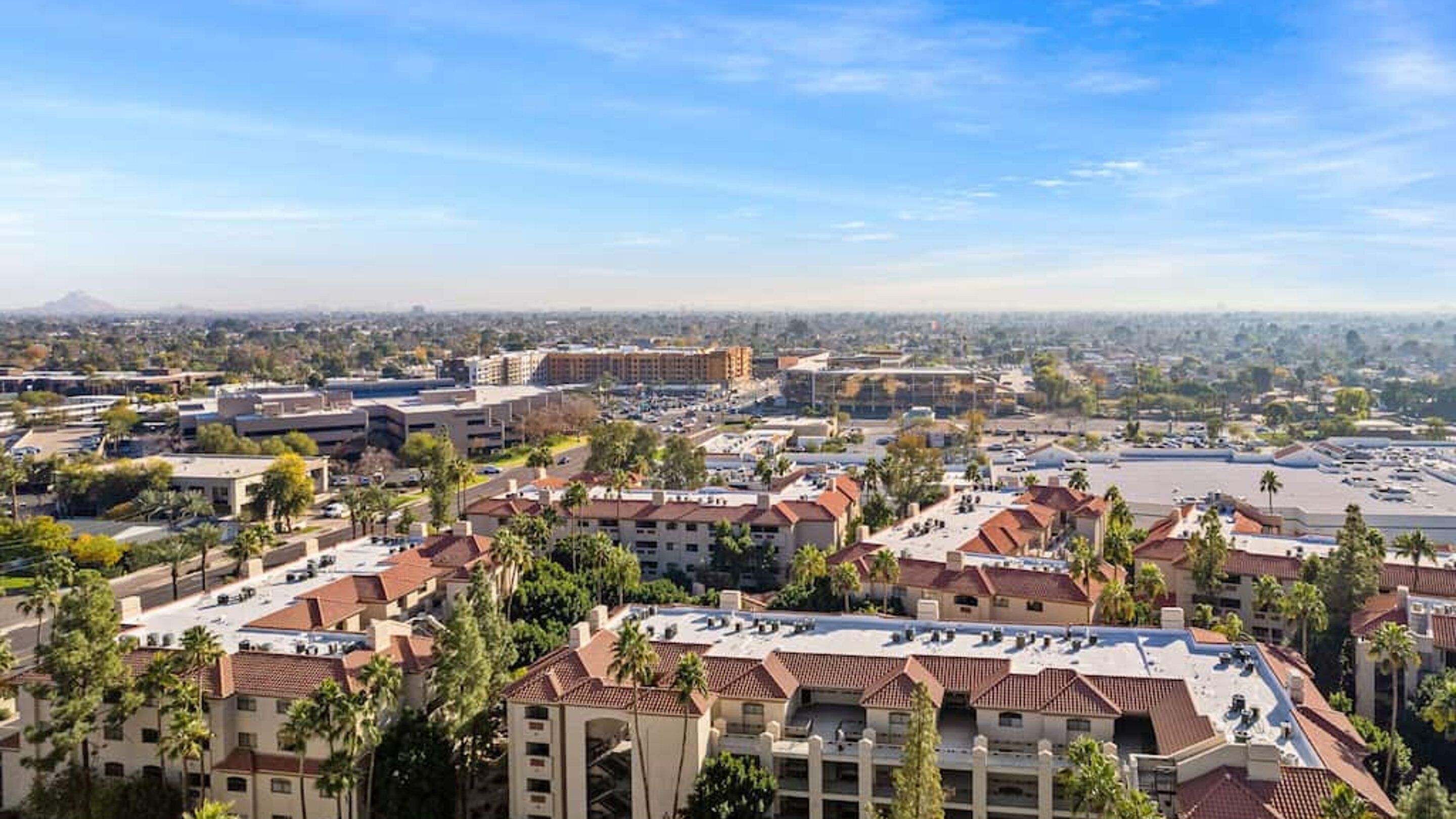 A residential area with multiple apartment buildings, some with red-tiled roofs and others with flat roofs. There are palm trees and a large parking lot visible.