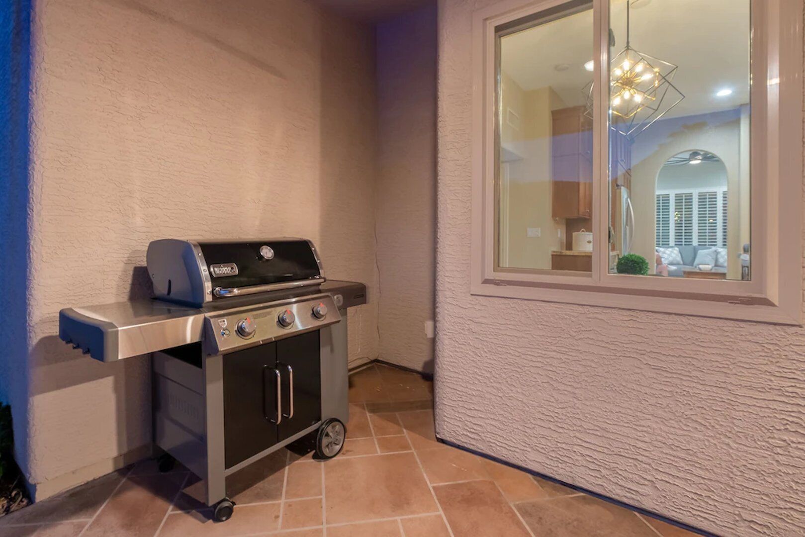 A portable grill with four burners and a side burner, placed against a textured wall with a window reflecting interior lighting.