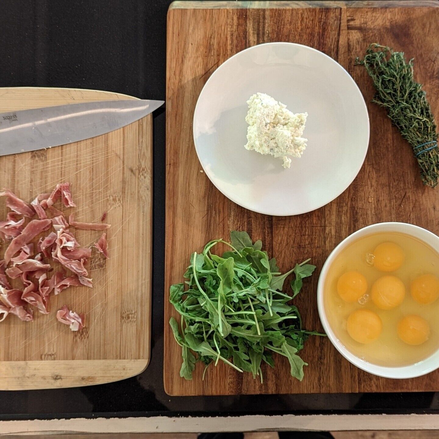 A wooden cutting board with a knife, chopped meat, fresh greens, a bowl of eggs, and a plate with a white substance.