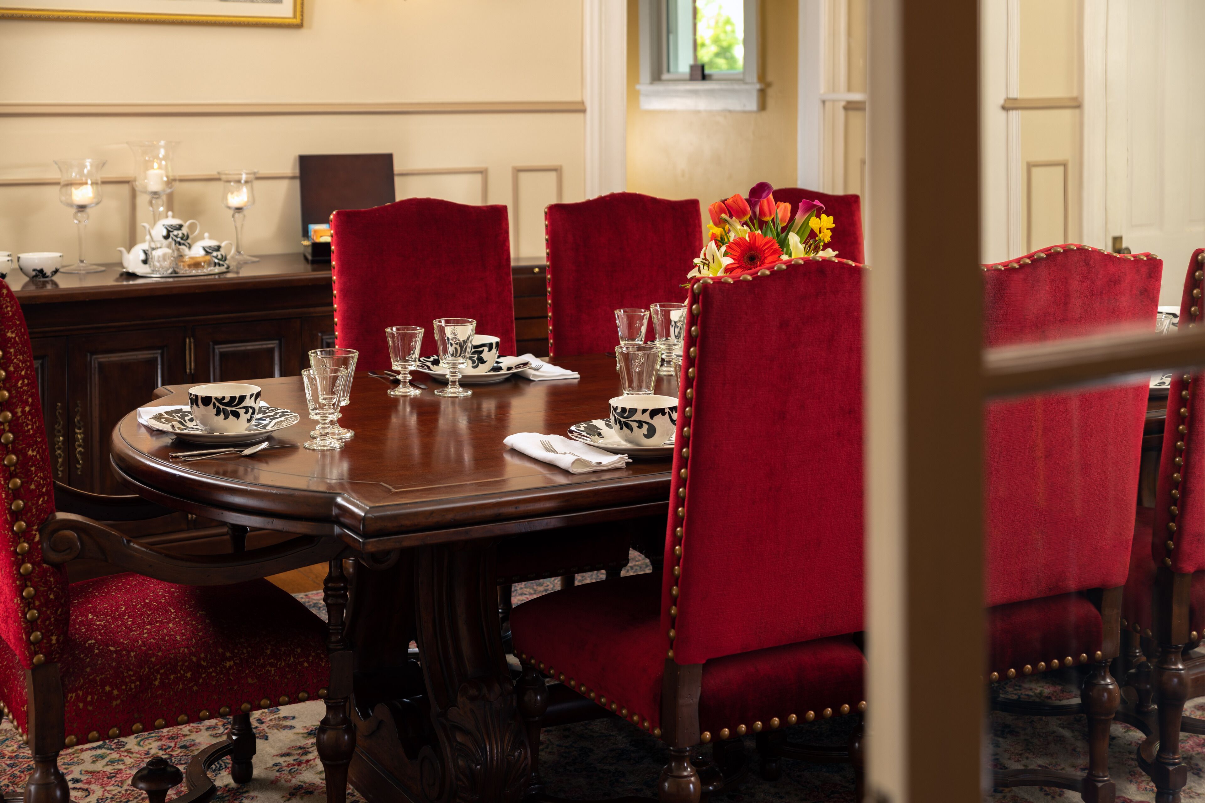 A formal dining room with a wooden table set for four, red upholstered chairs, and a vase of flowers.
