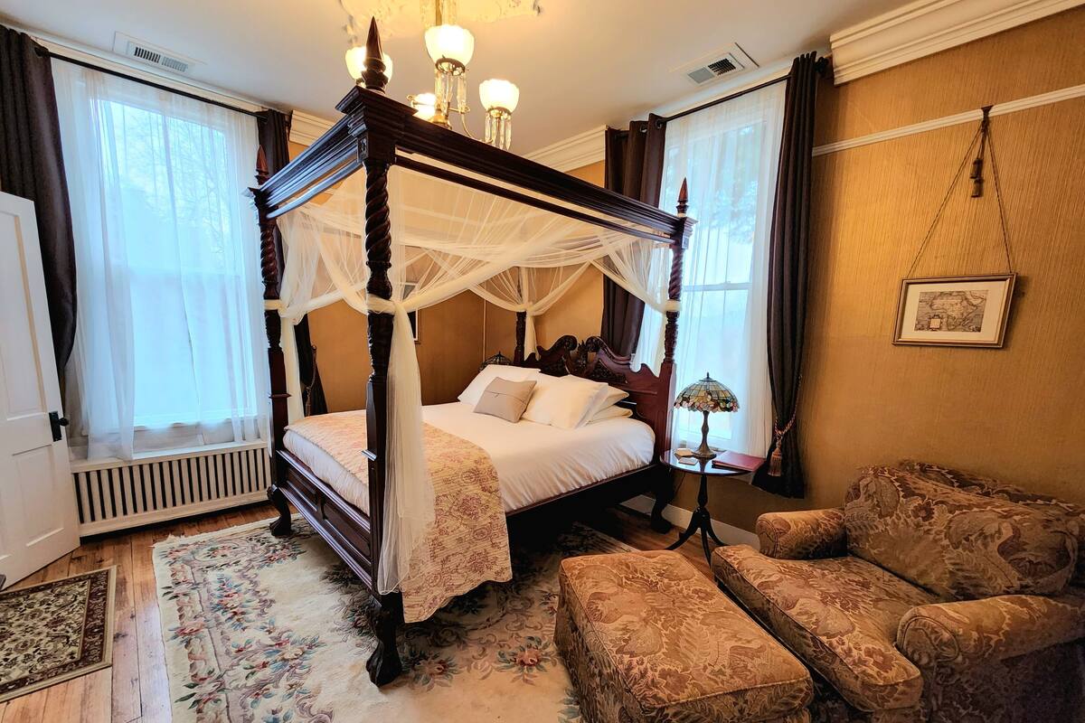 Una cama con dosel con dos columnas y dosel, con una colcha estampada de motivos florales, un reposapiés a juego, una mesita de noche con lámpara y un sillón con estampado.