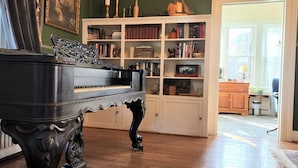 A grand piano in a room with a wooden floor, a bookshelf filled with books and decorative items, and a large window with a view of the outside. - South Court Inn (Luray)