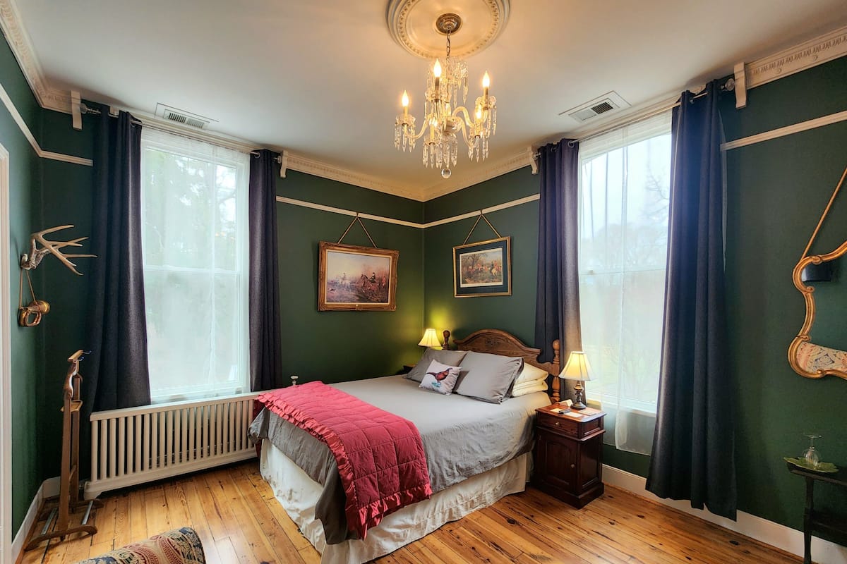 Un dormitorio con suelo de madera, una cama con manta roja, dos lámparas de noche, un candelabro y dos cuadros en la pared.
