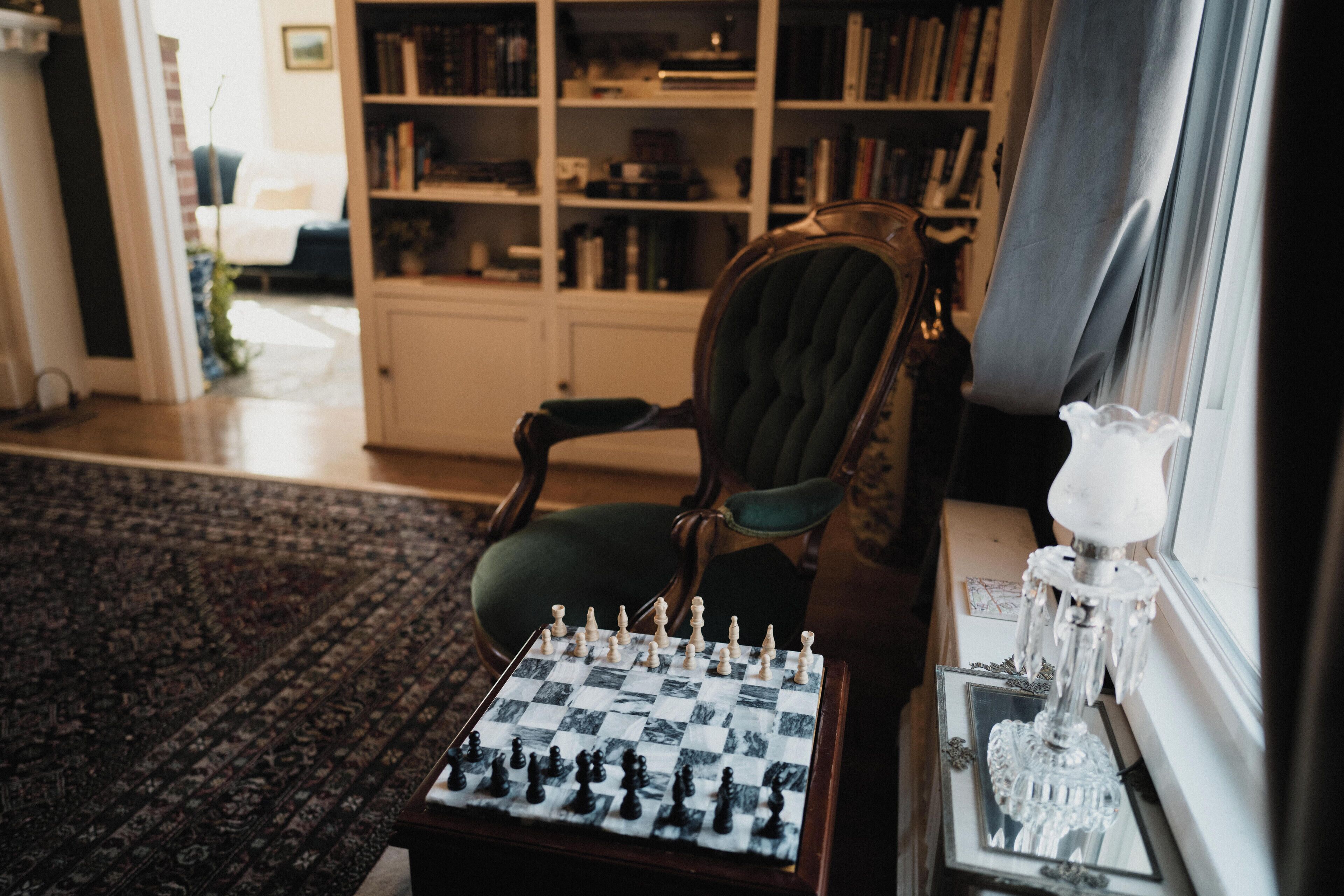 A chessboard on a table, a chair, a bookshelf, and a window with a lamp.