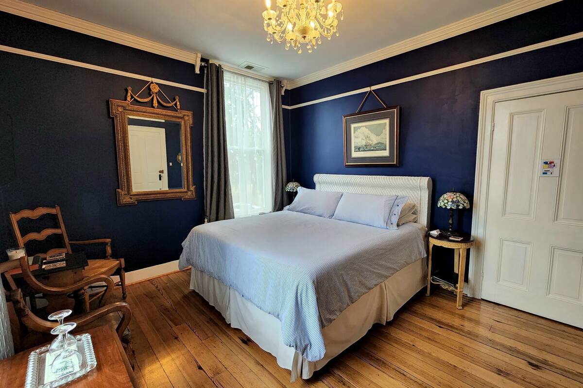 Un dormitorio con una cama grande, una silla de madera, una mesita de noche con una lámpara, un candelabro, un espejo y una puerta.