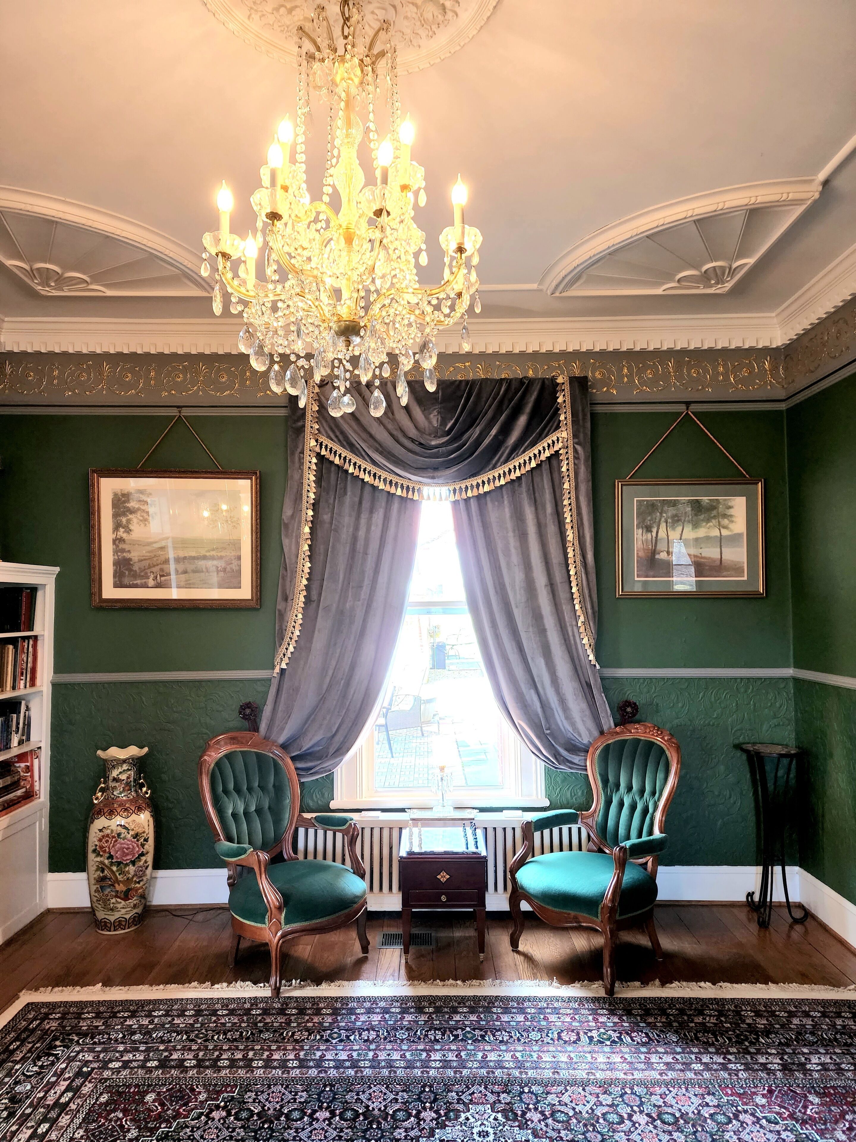 A room with a chandelier, two green upholstered chairs, a patterned rug, and framed pictures on the walls.