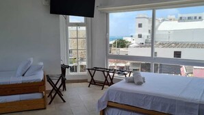 A room with two beds, a TV mounted on the wall, and a large window offering a view of buildings and the sea.