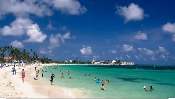 A beach with clear turquoise water, palm trees, and buildings in the distance.