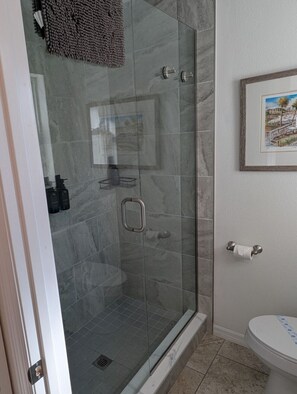 Une douche fermée par des parois de verre, avec un sol en marbre, un tableau encadré au mur et des toilettes à motifs bleus.