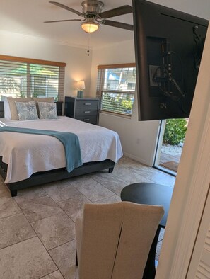Une chambre à coucher comprenant un lit, un bureau, une chaise, un ventilateur de plafond et un téléviseur fixé au mur.