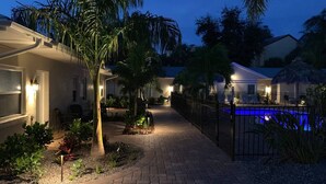A well-lit pathway leading to a pool area with a fence, surrounded by palm trees and a brick walkway. - #7-TwinPalms-King Studio Suite (Siesta Key)
