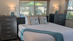 A bedroom with a bed, two bedside tables, a nightstand, a window with blinds, and a view of greenery outside. - #7-TwinPalms-King Studio Suite (Siesta Key)