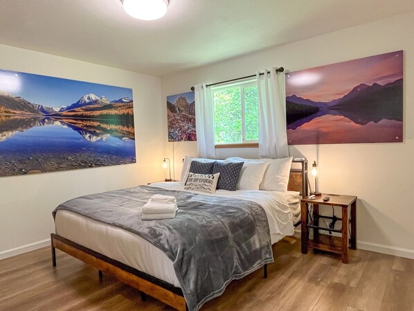 A bedroom with a bed, two bedside tables, a window with curtains, and three large landscape paintings on the walls. - Modern Cabin - 12 Minutes to Glacier! (Hungry Horse)