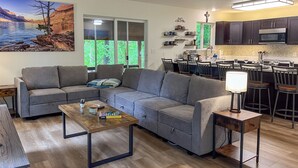 A modern living room with a gray sectional sofa, a wooden coffee table, and a dining area with bar stools. A kitchen with dark cabinets and a microwave is visible in the background. - Modern Cabin - 12 Minutes to Glacier! (Hungry Horse)