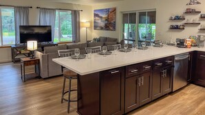 A modern kitchen with a central island, dark cabinetry, and a dining area with a sofa and TV. - Modern Cabin - 12 Minutes to Glacier! (Hungry Horse)