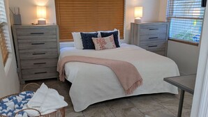 Quarto com cama, mesas de cabeceira, escrivaninha, ventoinha no teto e janela com persianas.