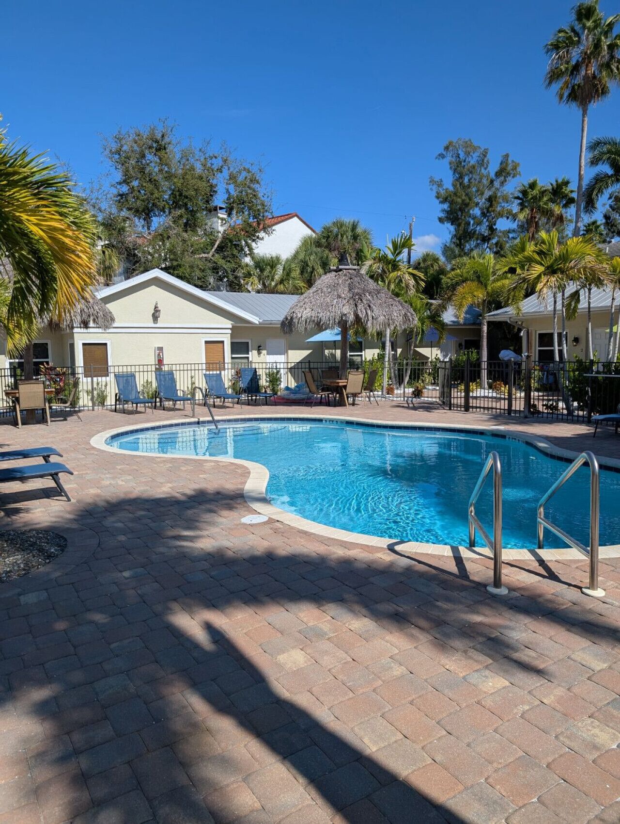 A swimming pool with lounge chairs and a thatched umbrella, surrounded by palm trees and a building with a white roof.