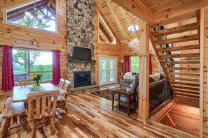 A wooden cabin interior with a stone fireplace, dining area, and a staircase.