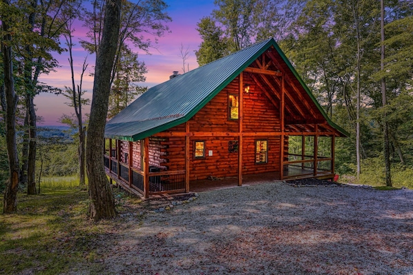 A rustic cabin with a sloped roof, surrounded by trees and a gravel driveway.