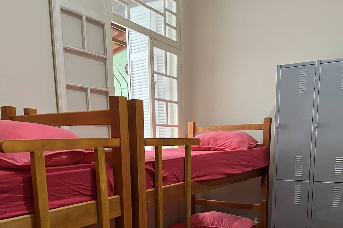 Une chambre avec un lit superposé en bois et une literie rose, une fenêtre avec des volets blancs et un casier métallique gris.