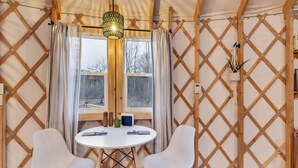 A cozy room with a round table, two white chairs, a hanging light fixture, and a window with curtains.