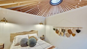 A bedroom with a wooden ceiling, a bed with a wooden headboard, two bedside lamps, and a wall-mounted shelf with decorative items.