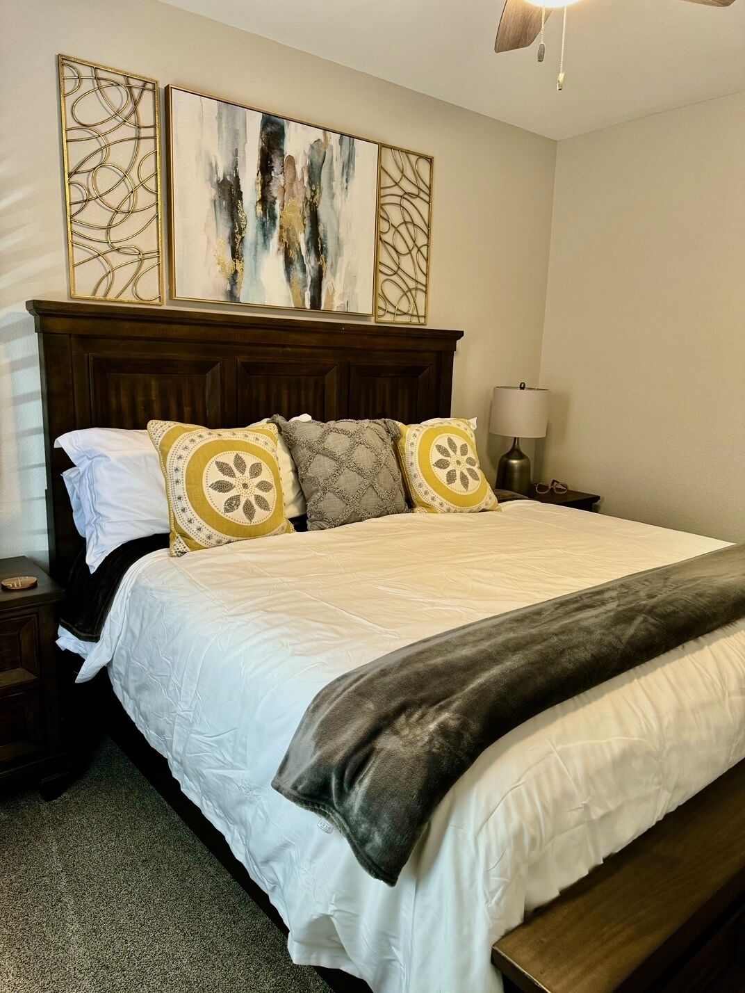 A neatly made bed with a white comforter and decorative pillows, a wooden headboard, and a nightstand with a lamp.