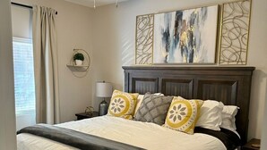Une chambre à coucher avec un lit en bois foncé, du linge de lit blanc, un ventilateur de plafond et des œuvres d’art abstraites accrochées au mur.