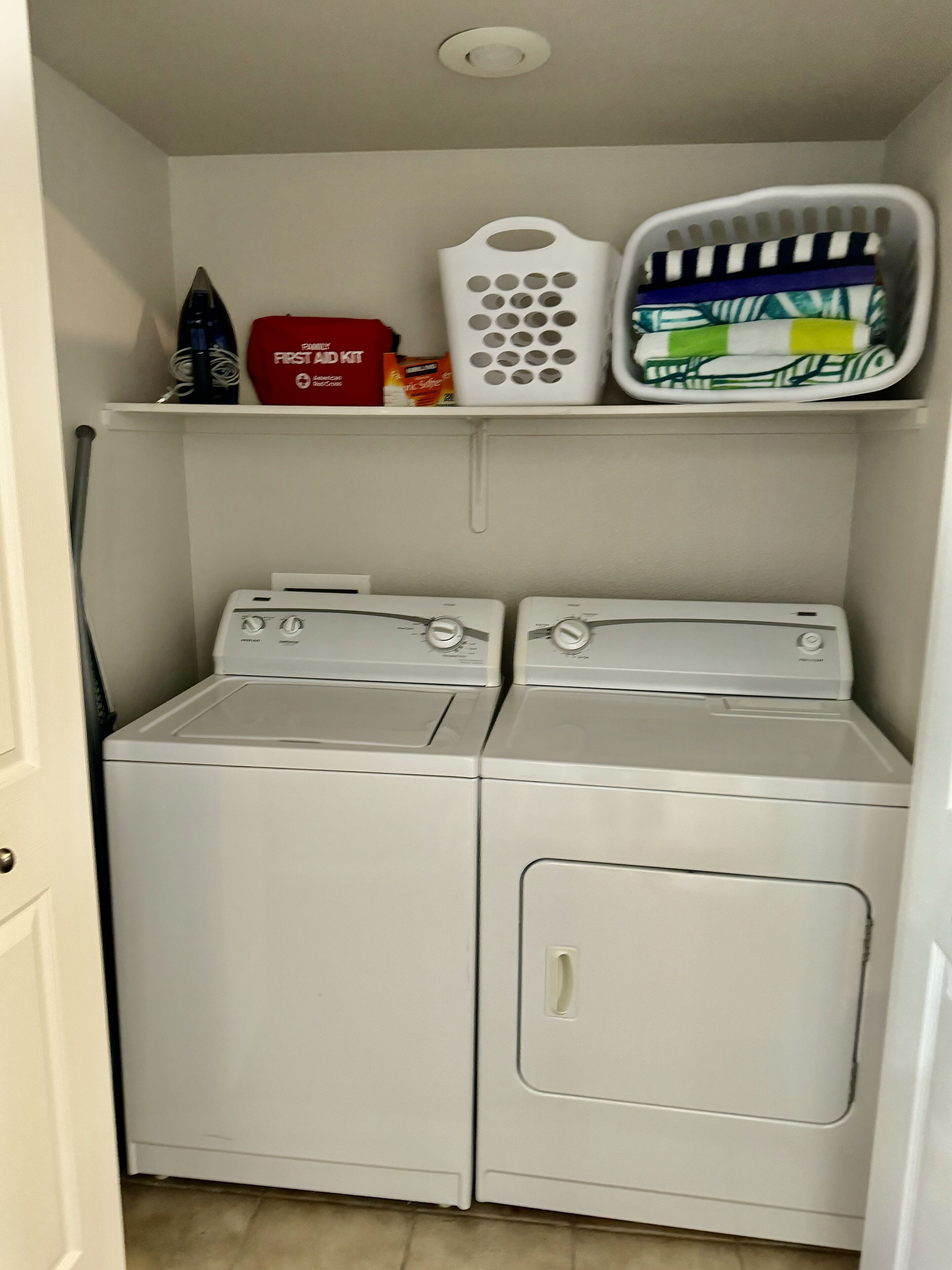 A laundry room with two front-loading washing machines and a shelf above containing a red box, a white basket, and a folded green and white blanket.
