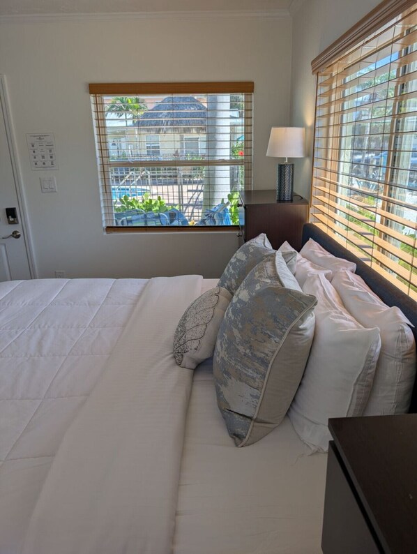 A bedroom with a bed, pillows, a nightstand, a lamp, and a window with blinds. - #8-TwinPalms-King Studio Suite (Siesta Key)