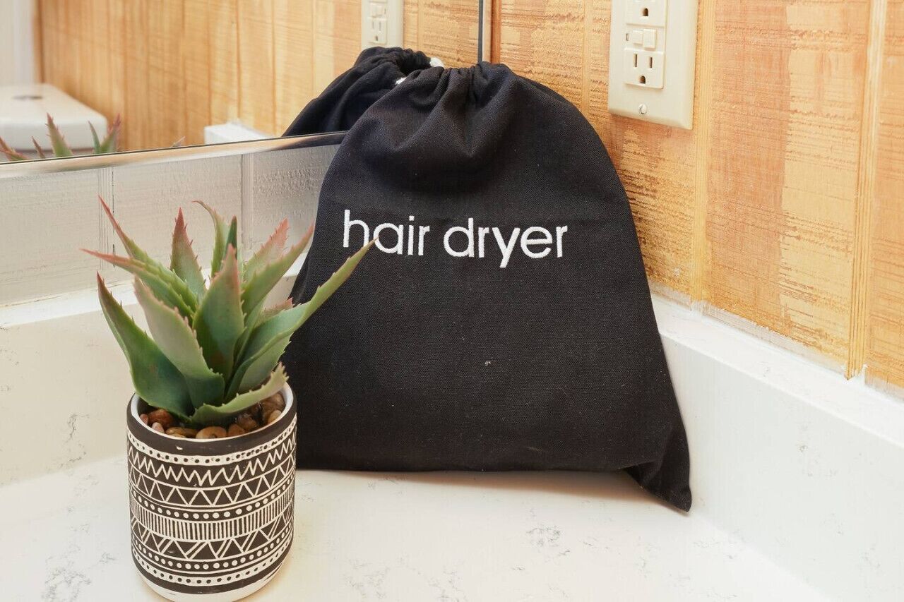 A black drawstring bag with "hair dryer" written on it, placed next to a potted plant on a bathroom counter.