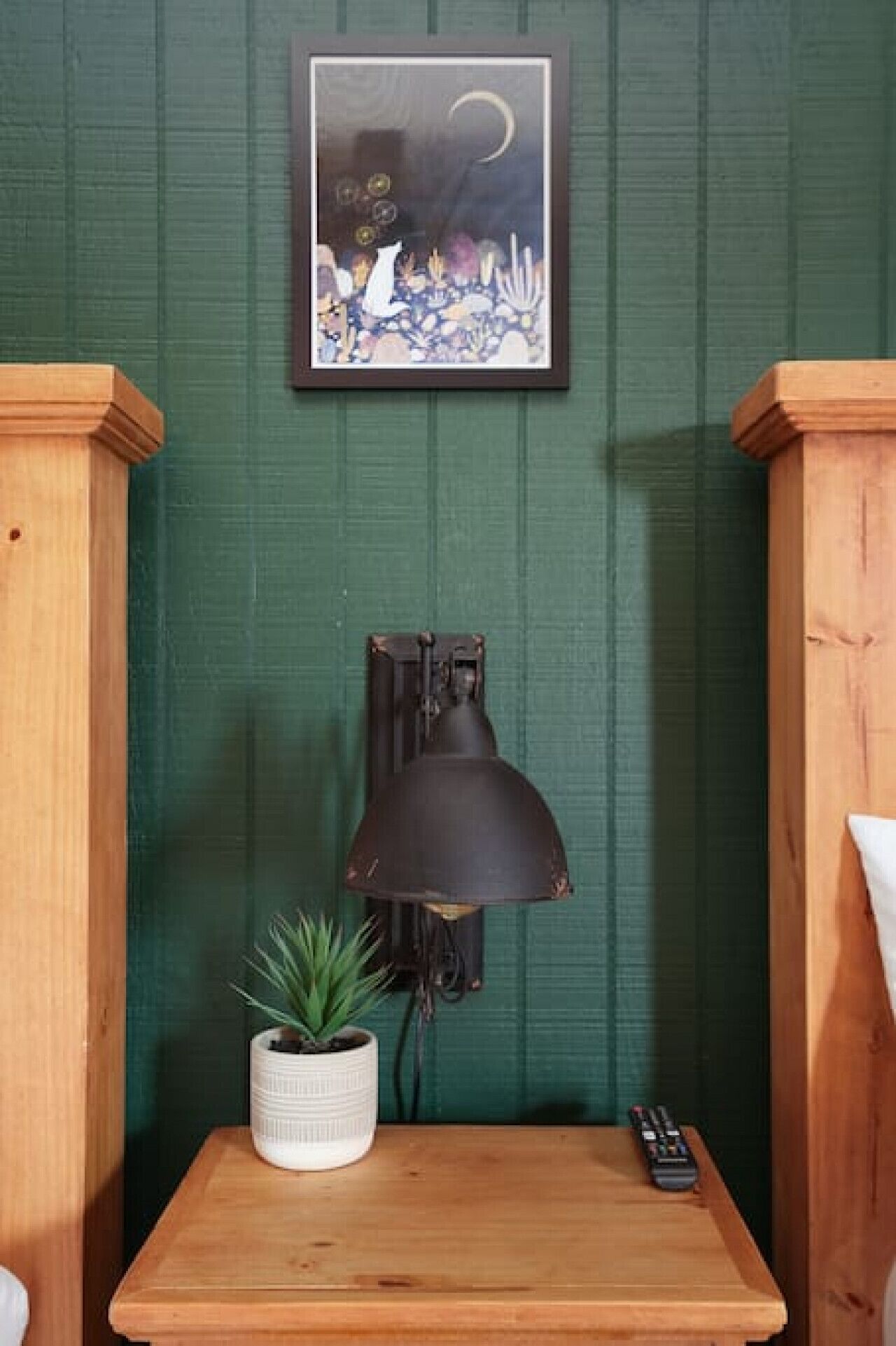 A room with a green wall, a framed artwork, a desk with a lamp, a potted plant, and a remote control.
