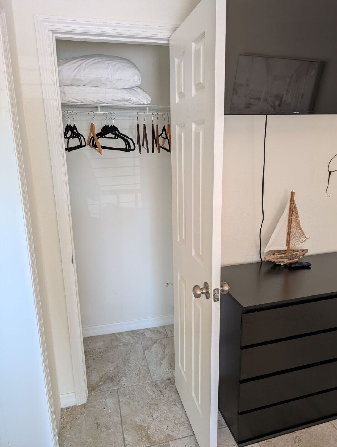 Ein Zimmer mit Kleiderschrank, gefüllt mit Kleidung, und einem Bett mit weißen Laken. Eine schwarze Kommode mit Schubladen und ein kleines Schiffmodell darauf.