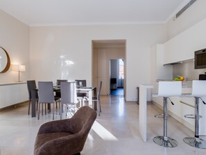 Un saló modern amb una butaca marró, una zona de menjador amb taula i cadires, i una cuina amb un televisor i un plat de fruita.