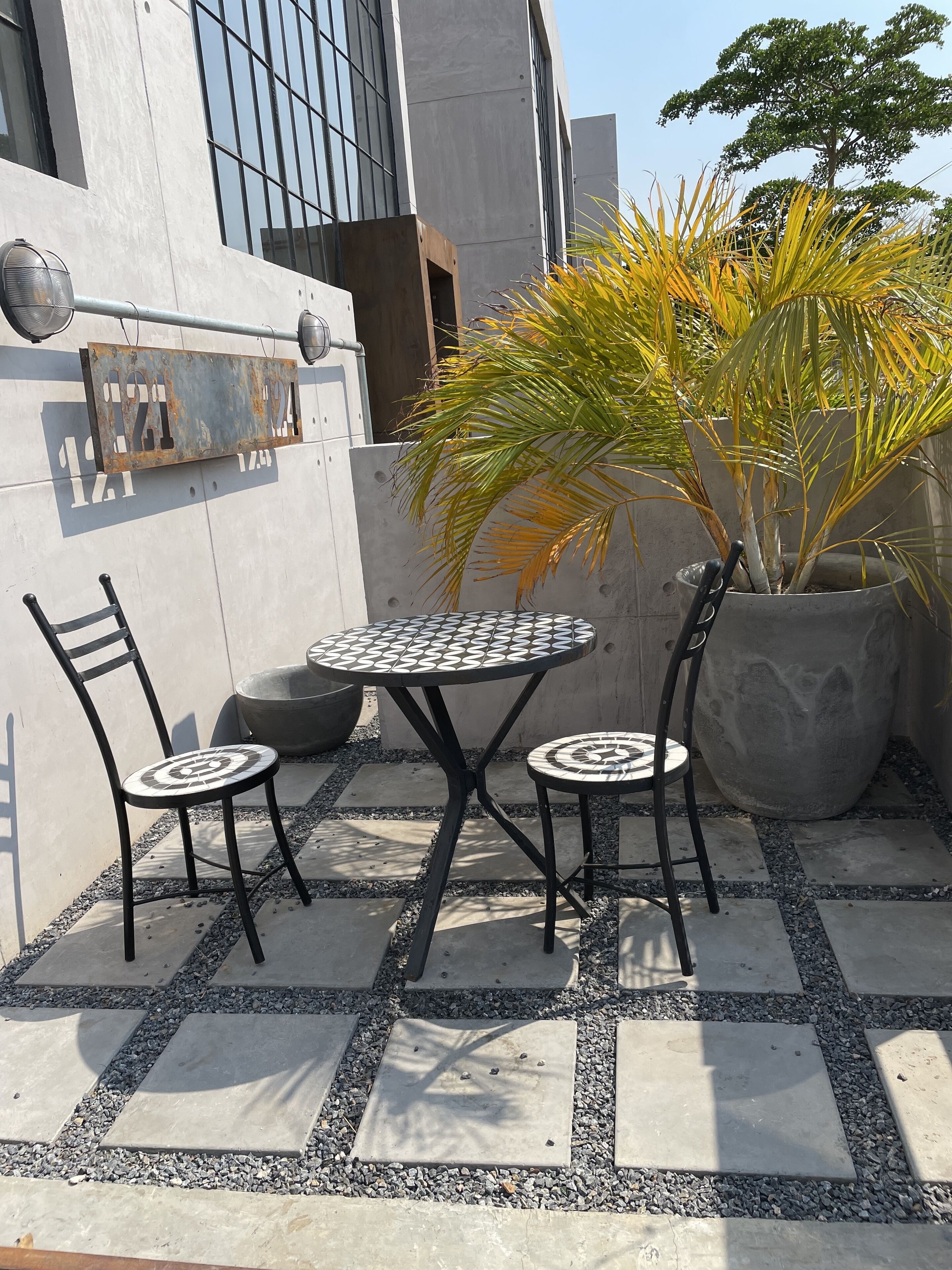 Een buiten zitgedeelte met twee metalen stoelen en een ronde tafel, omgeven door een vloer met een patroon en een potplant.