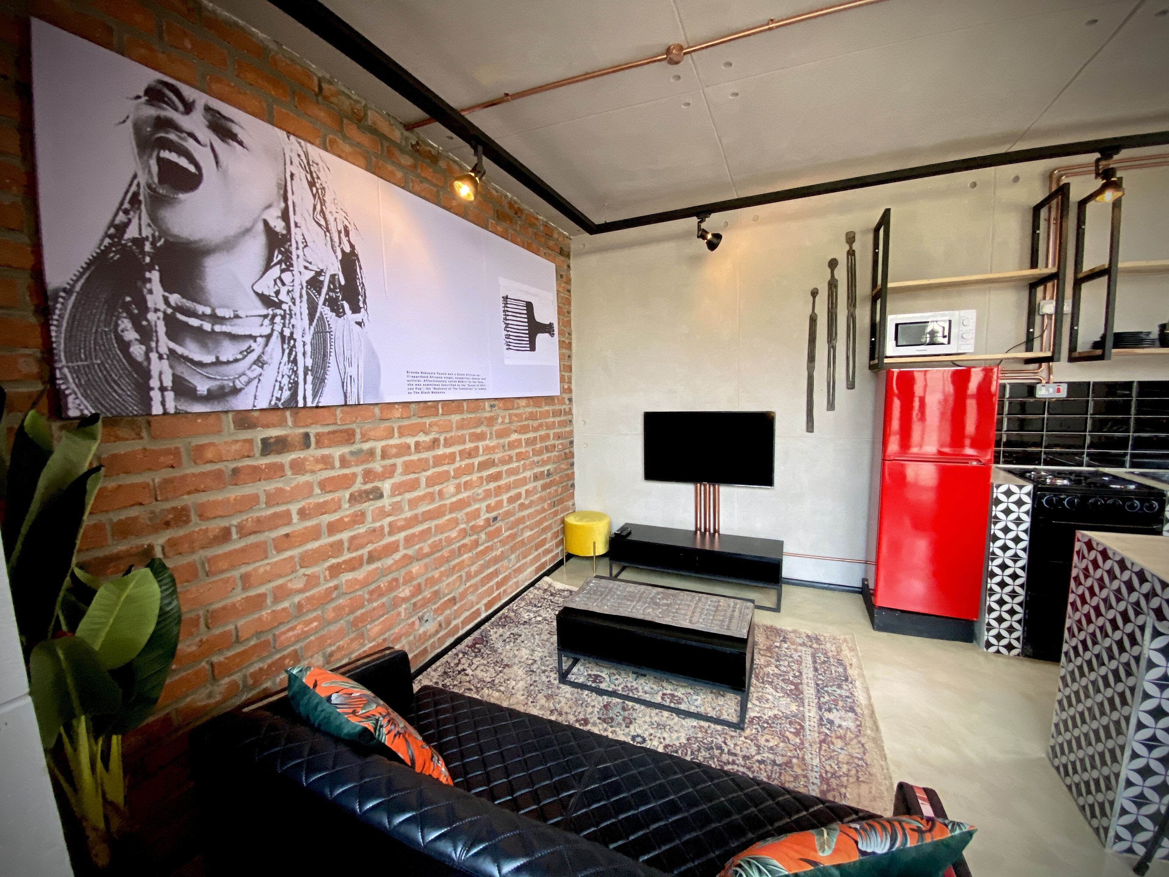 Un espacio habitable moderno con pared de ladrillo, televisor de pantalla plana, refrigerador rojo y sofá negro con cojines estampados.